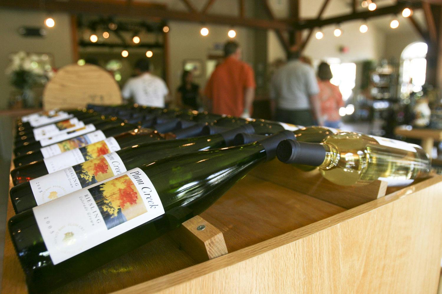 Atop a wooden shelf, there are green and glass bottles of wine lined up. In the distance people listen into a wine tasting at Plum Creek Winery in Palisade.