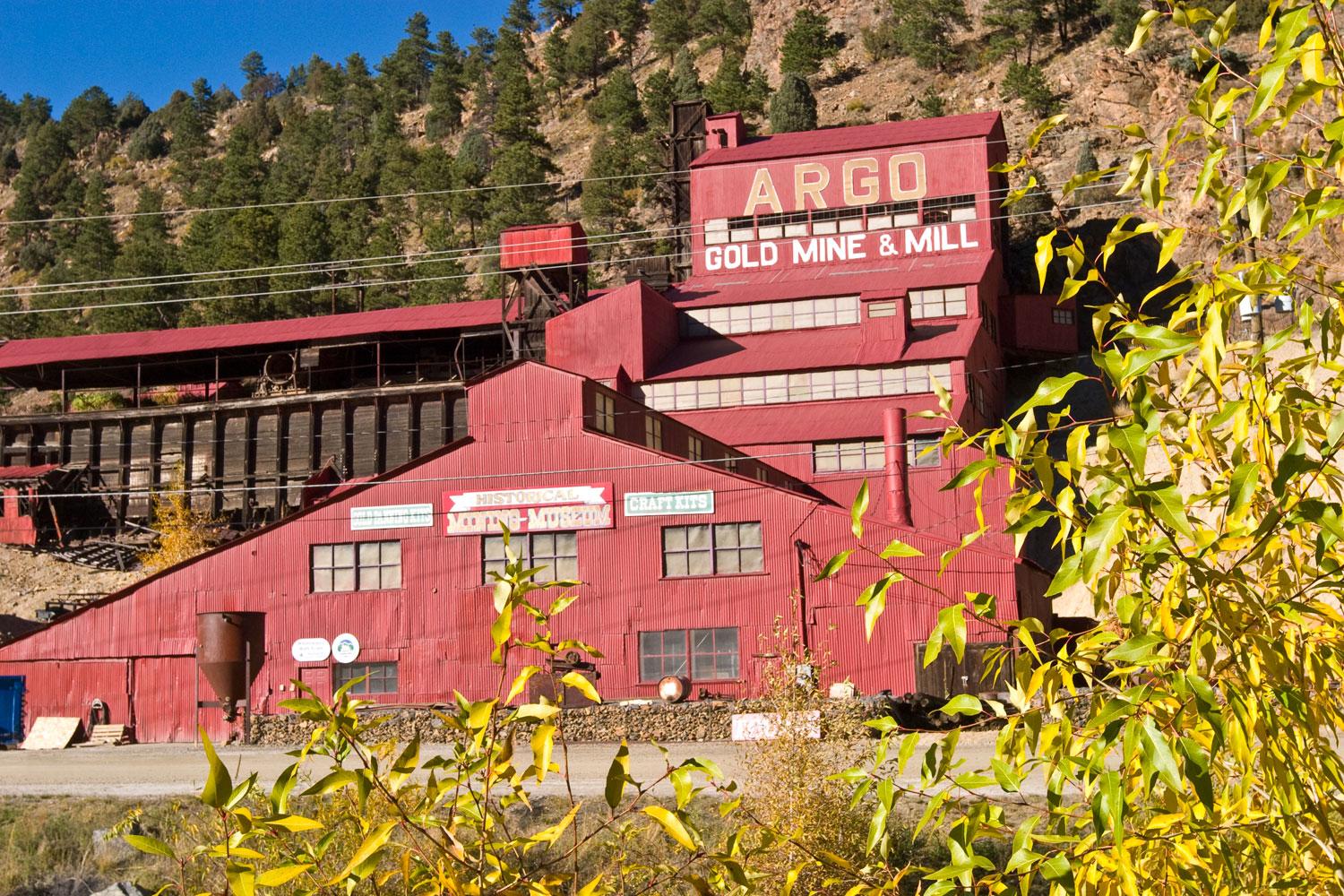 Bright-red industrial buildings stack up against a mountainside; a sign is painted on that reads "ARGO Gold Mine & Mill"