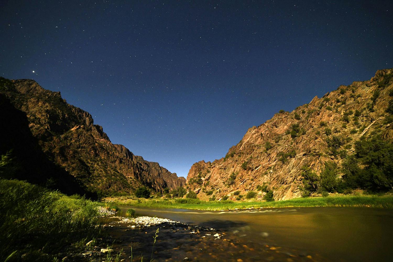 The deep-blue night sky sits above rock-walled canyons with a river with green-grass covered riverbanks.