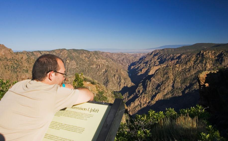 A person leans over an interpretive sign and gazes into the distance at the striped walls of the Black Canyon of the Gunnison National Park near Montrose, Colorado