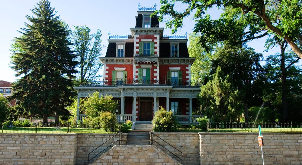 A three-story mansion pained red, with white brick accents, green shutters and a black mansard roof sits behind a brick wall and manicured lawn