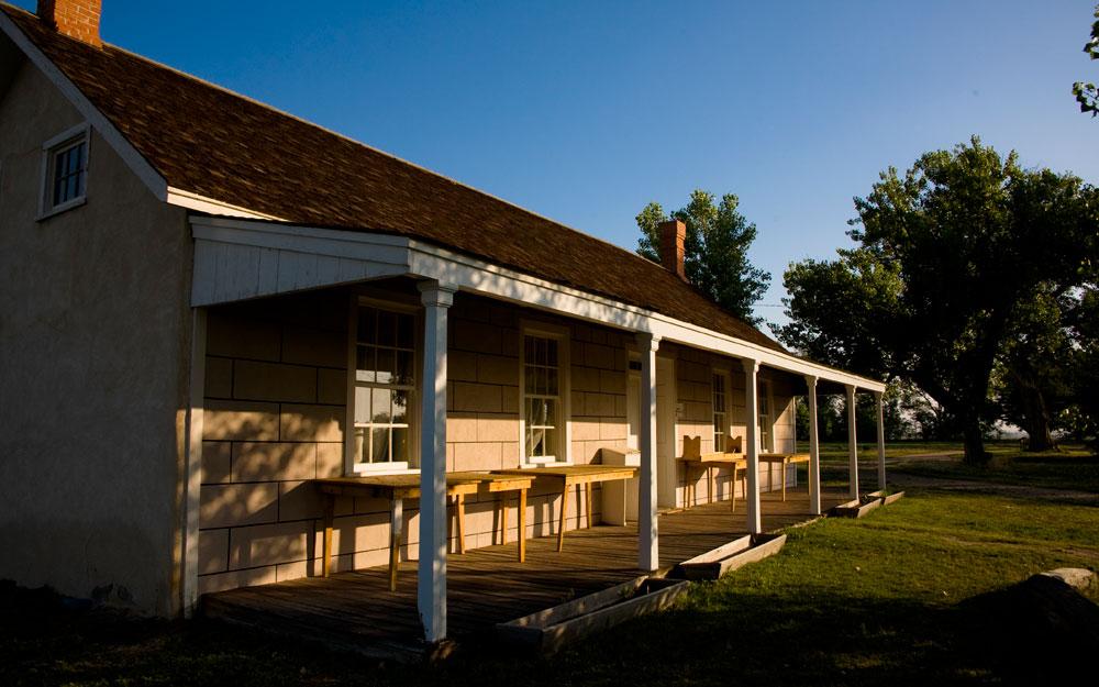 In Boggsville, Colorado, a lone building with a low-roofed porch, featuring several plain wooded tables, is set among several mature trees on a grassy lawn.