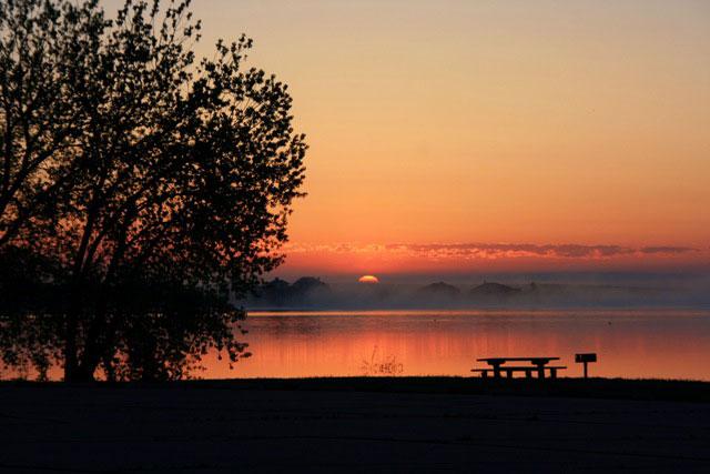 A fiery orange sunset descends over a tree, picnic table and lake in an otherwise empty park