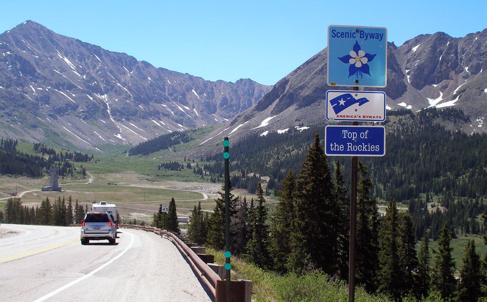 A car and RV drive down the curve of a Colorado scenic byway. Ahead, winding paths lead through a valley between two peaks with pockets of snow.