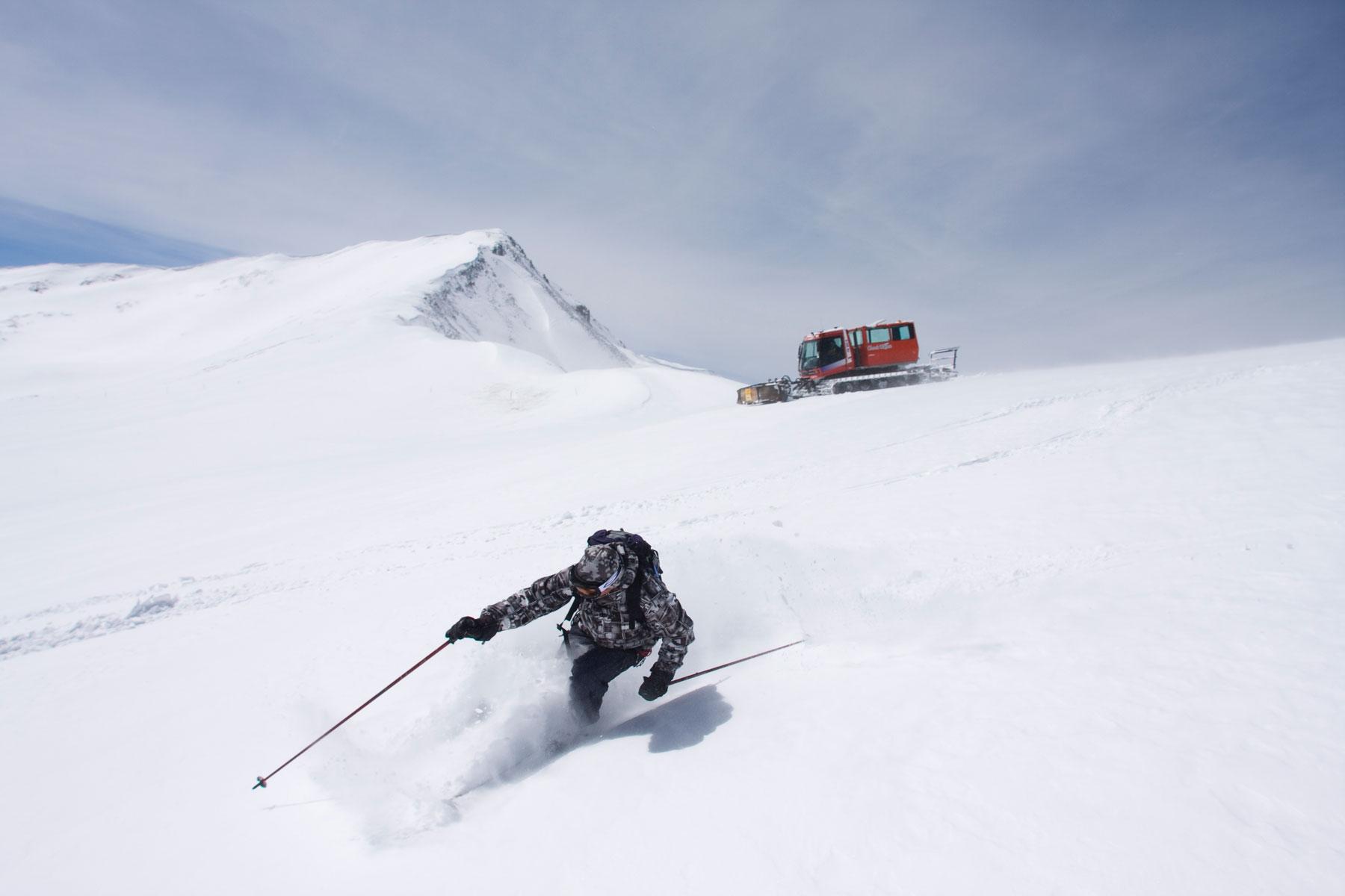 At the top of a very snowy, very powdery hill, a snowcat is parked. In the foreground, a skier plows through kneed deep powder with poles outstretched