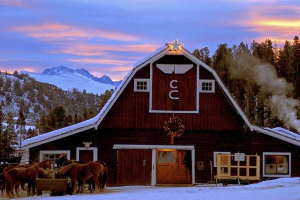 A broad, white-trimmed, red-faced barn sits comfortably in front of distant snow-capped mountains in Granby. On the top of the barn is a small, golden glowing star, underneath which are two "c"s. This is the C Lazy U Ranch. In front of the barn is a small group of brown horses eating from a trough.