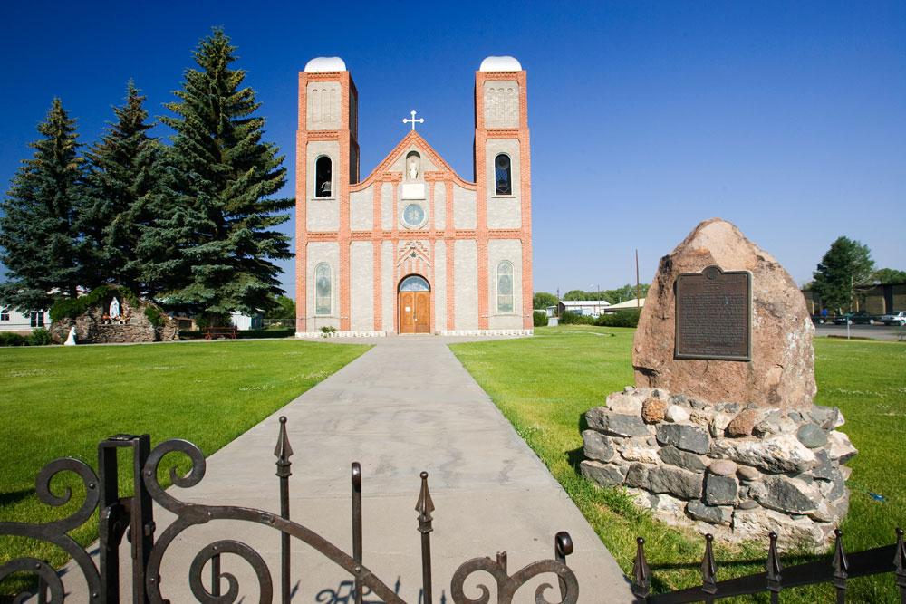 A tall church, featuring two towers with red, decorative brickwork, sits on an immaculate green lawn in Conejos, Colorado.