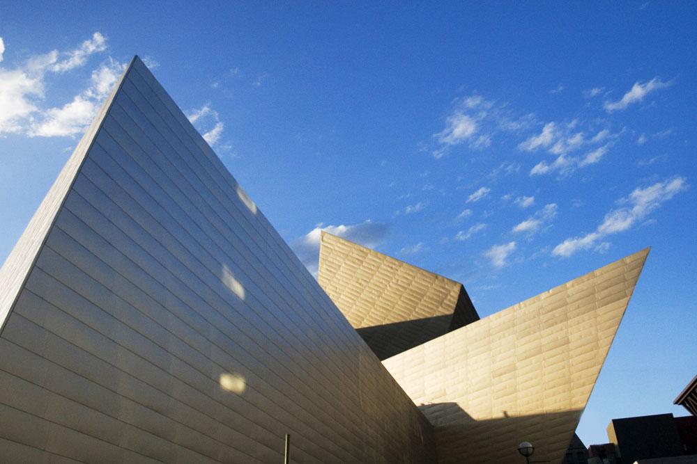 A mostly-clear, bluebird sky frames the pointed, triangle-like roof design of the Denver Art Museum in Denver, Colorado. 