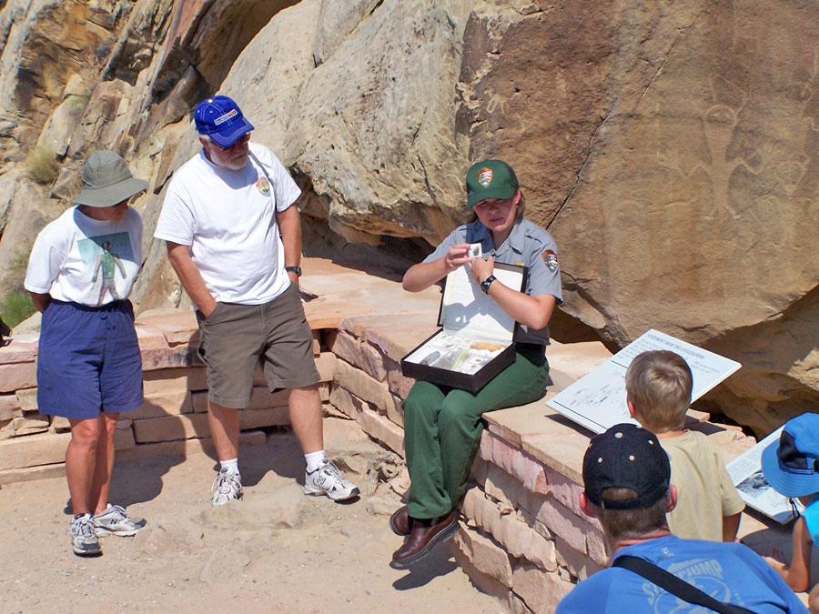 A national park ranger in green pants and a green hat showcases a box full of items to five tourists. She sits on a low rock wall with a rock with a pictograph on it behind her.