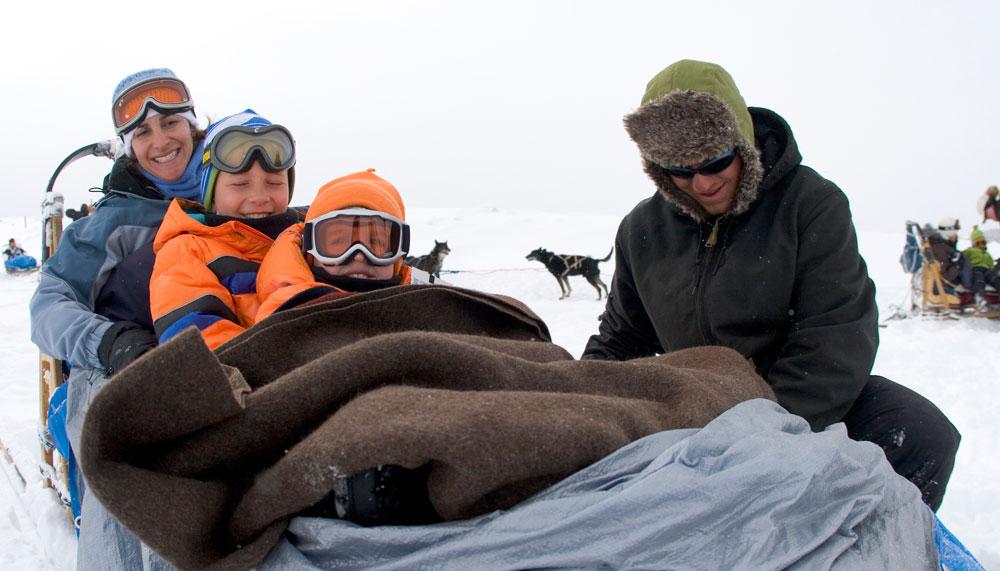 Three people sit in a dog sled while a musher secures their brown blanket. In the background two dogs face each other.