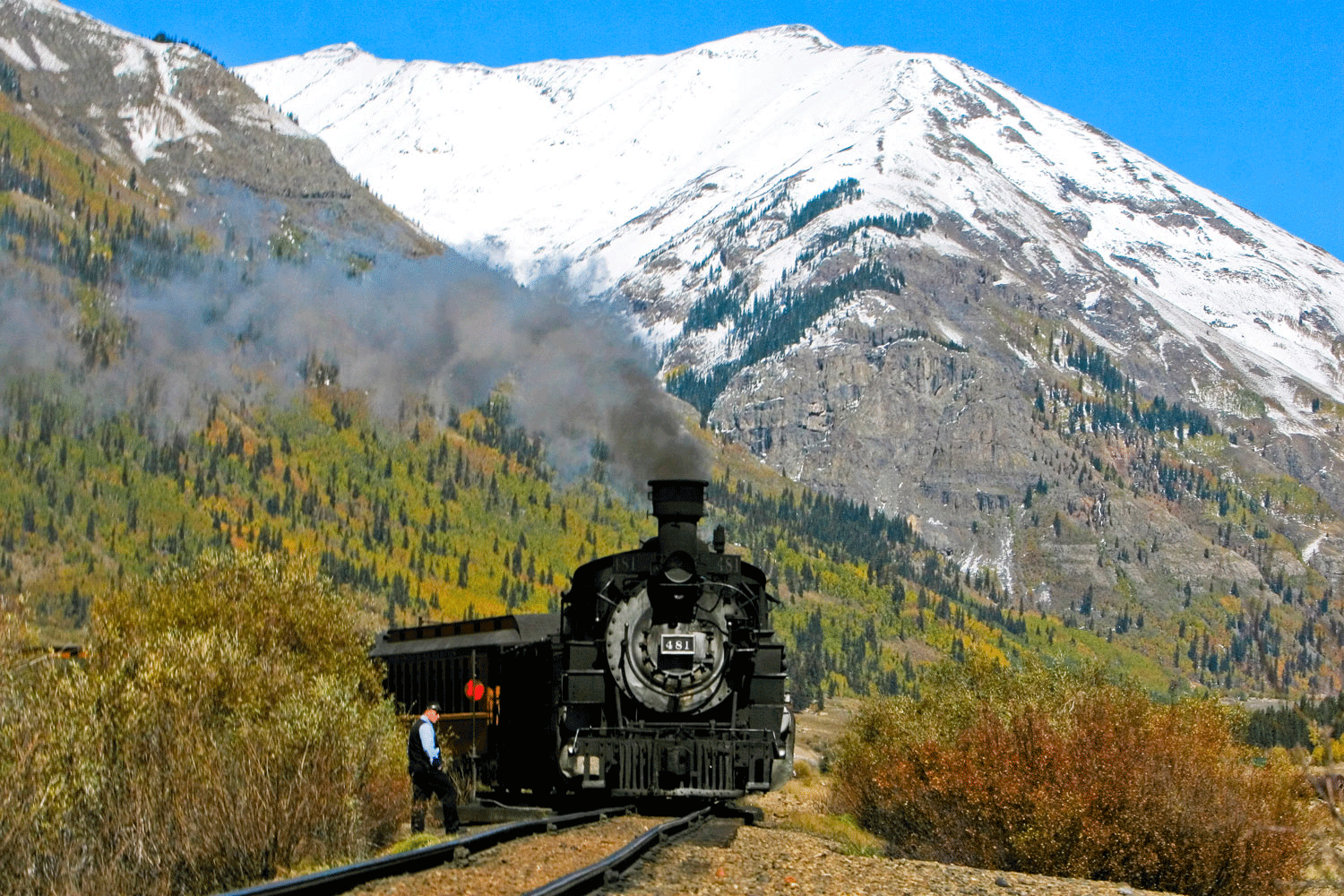 A train engineer stands to the side of a track as a black train engine with gray smoke billowing out its smokestack comes around the bend.