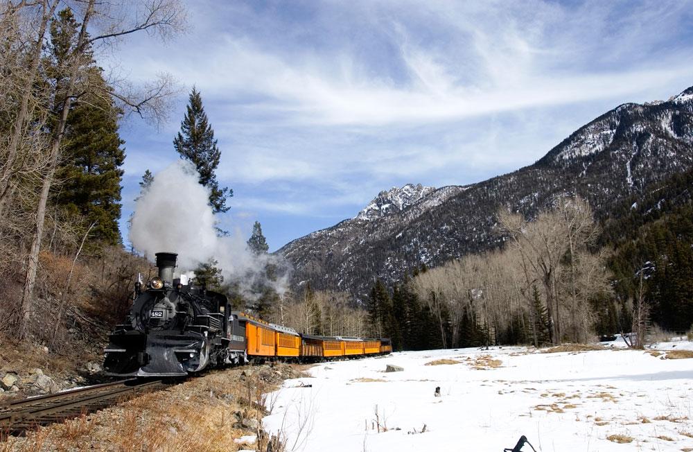 The Durango & Silverton Narrow Gauge Railroad hugs a mountainside curve with snow covering the ground and the mountains in the background.