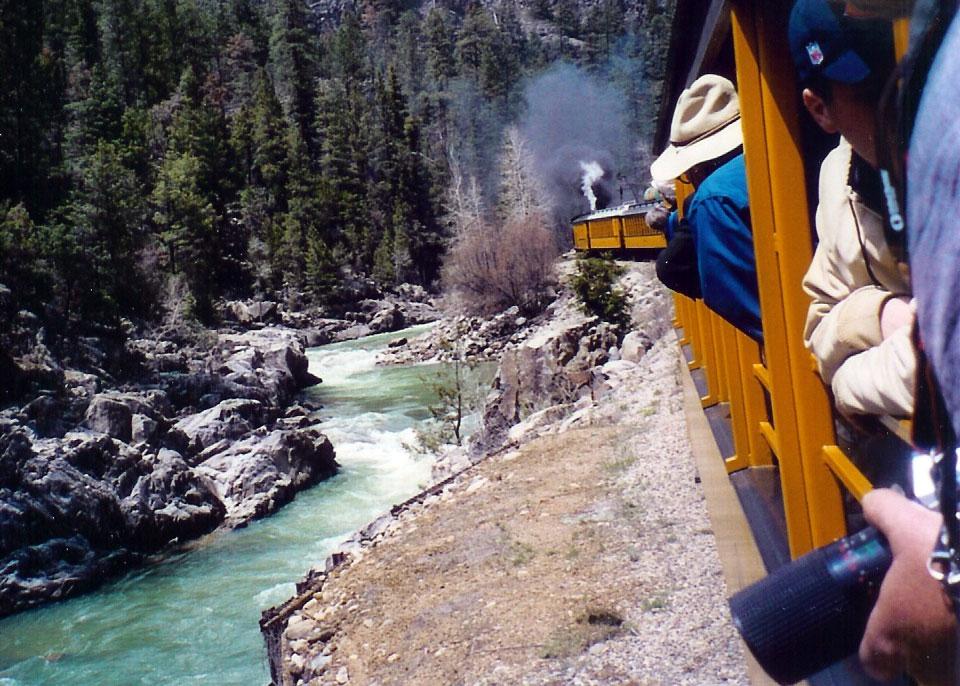 Passengers lean out the the side of an orange train car to take pictures and get a better view of the rush waters and forested peaks around them in Colorado.