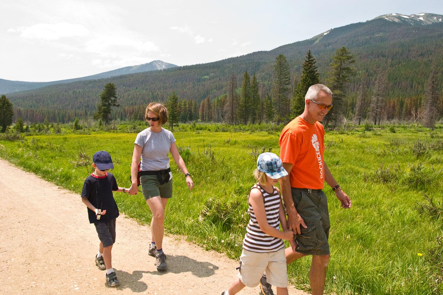 Two adults each walk besides a child along a dirt path through a meadow in Rocky Mountain National Park, Estes Park, Colorado. A pine tree forest and mountain peaks are in the distance.