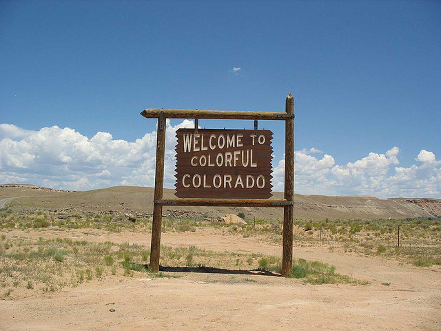 A large wooden sign is engraved with the words, "Welcome to Colorful Colorado."  Behind the sign are rugged Colorado mountains and a bluebird sky with clouds.