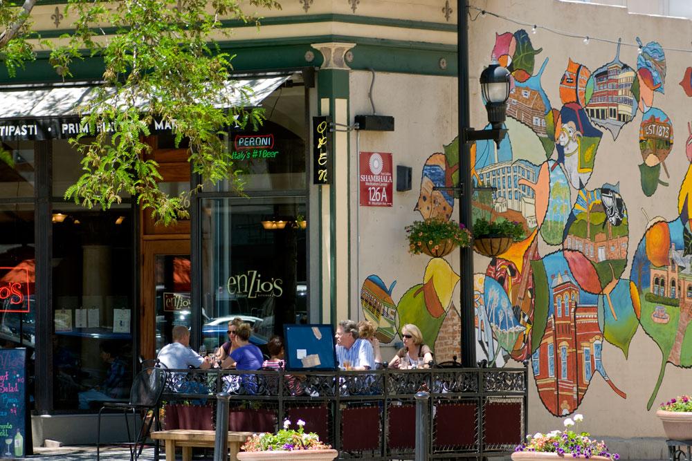 Diners sit at green tables outside a restaurant; on the side of the building, we see paintings of Fort Collins' buildings