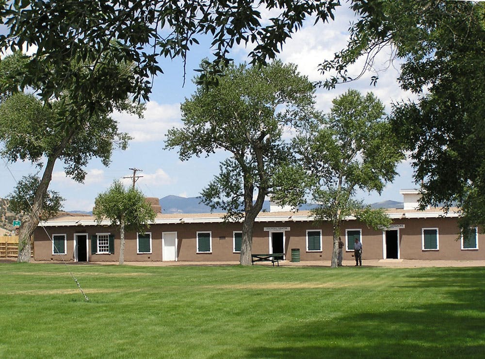 A long brown building with a flat roof is set among a well-maintained lawn and mature trees near Blanca.