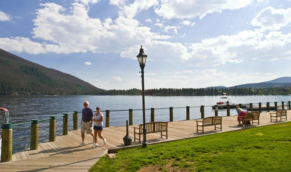 Two people walk along the wood boardwalk that runs along the blue waters of Grand Lake