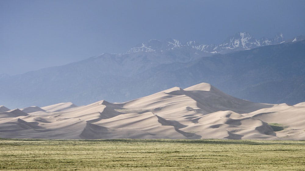 Just beyond a grassy meadow, mountains of windswept sand sit below a blue mountain range