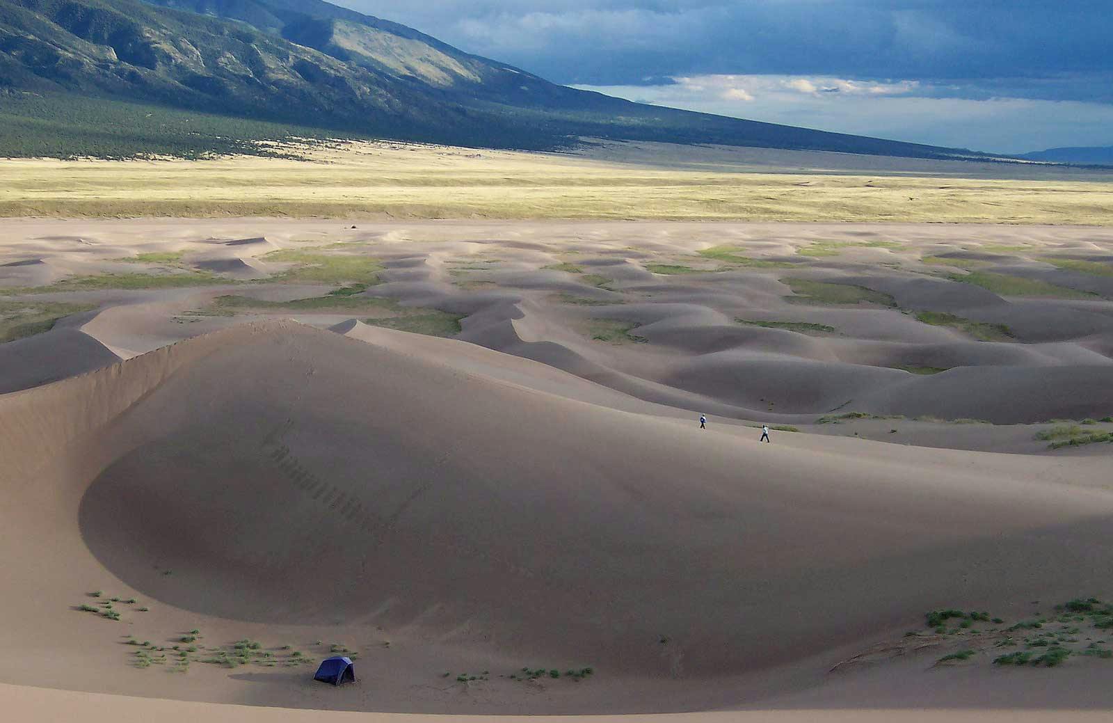 A blue tent rests next to a massive sand dune, with a field of sand dunes and mountains in the background