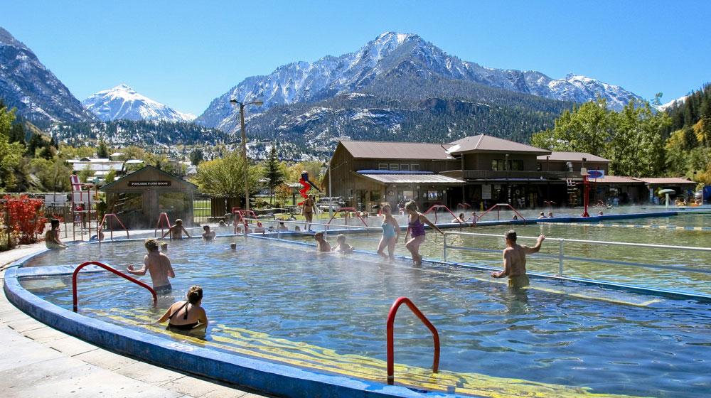 Visitors soak in the steamy waters of the Ouray Hot Springs Pool. In the distance are tall, forested, snow-capped mountain peaks.