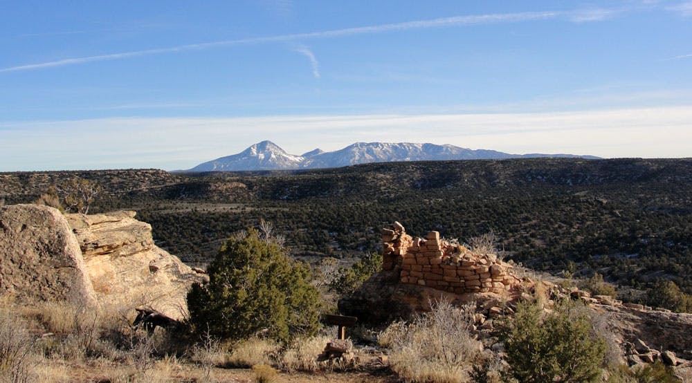 A crumbling brick structure sits high above a vast meadow that runs up to a snow-topped mountain peak at Colorado's Hovenweep National Monument.