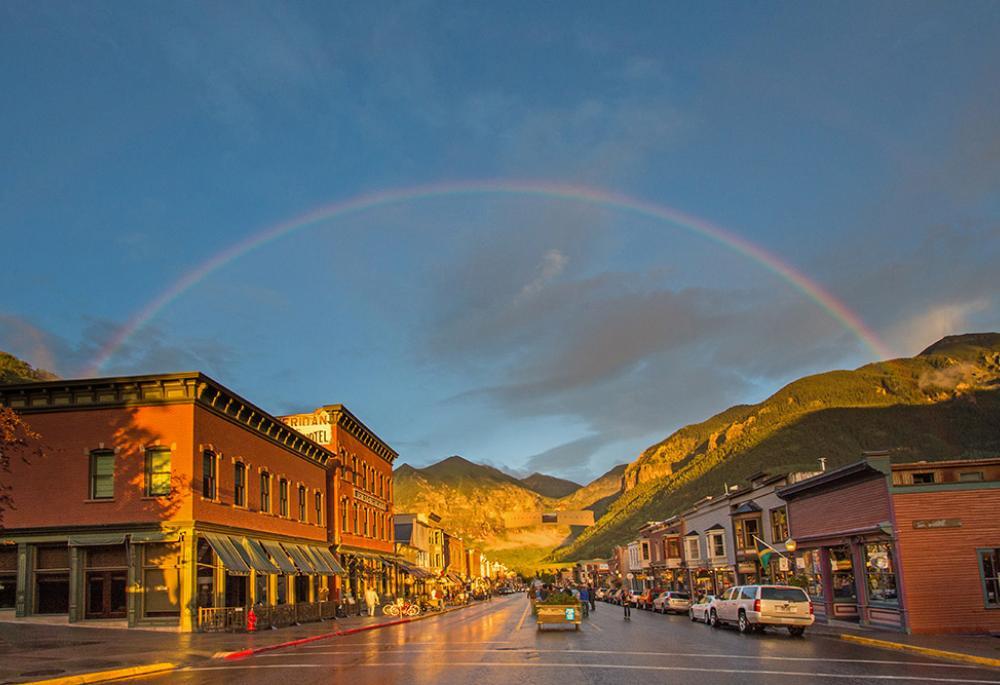 A rainbow arches above downtown Telluride as golden hour lights up the historic buildings and the mountain range.