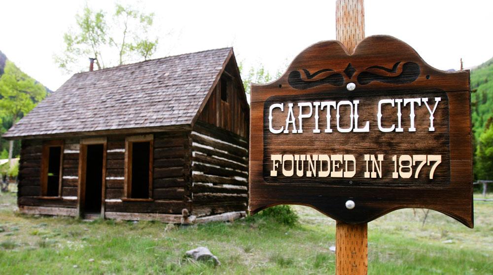 A wooden sign says Capitol City with a long cabin in the background and green grass on the ground.