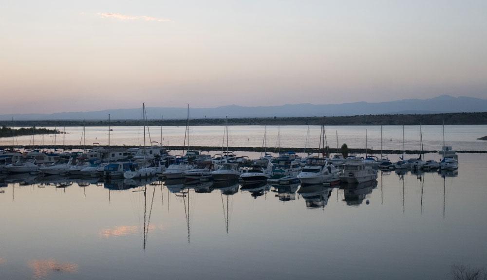 At dusk, a line of sailboats are docked along a lake's pier