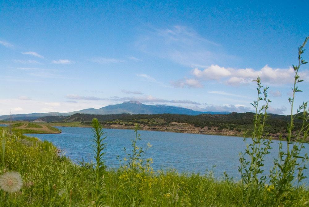 A lake with blue water is lined by green shrubs, and a mountain peak rises in the distance