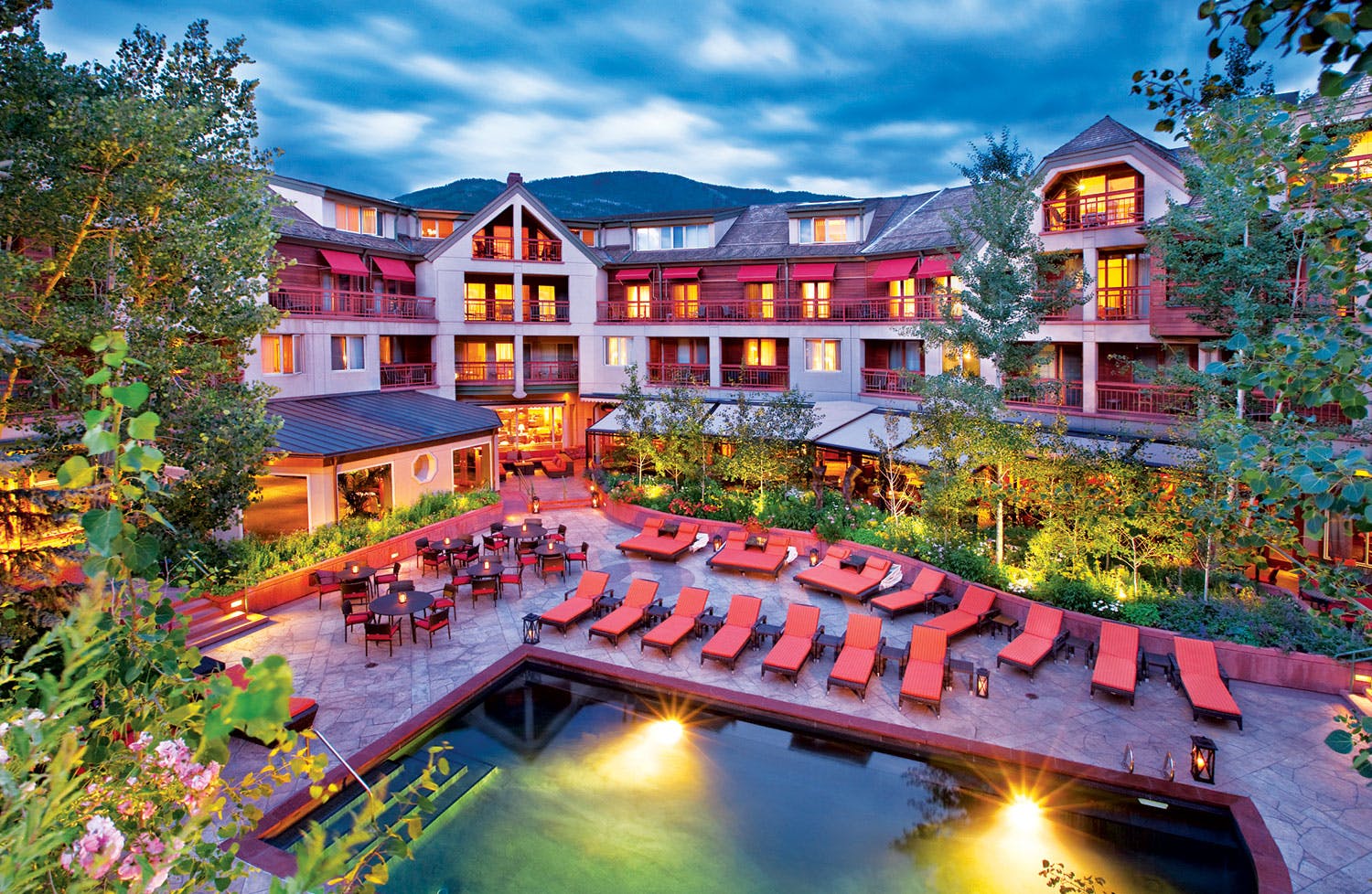 The Little Nell hotel in Aspen, a regal-looking building with the golden glow of lights through the various windows. The pool is cut off from the bottom half of the photo, and there are several orange chaise-longues line the path around the pool. The sky above is cloudy and gray, presumably after sunset.