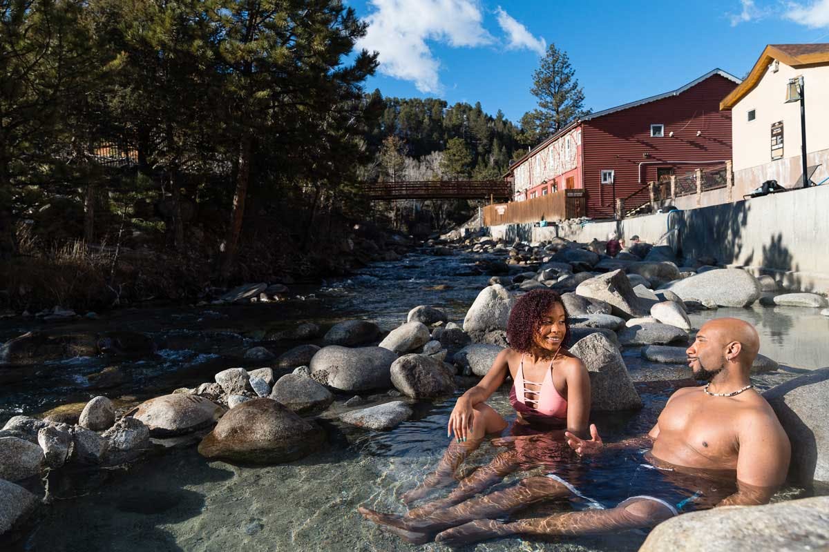 A couple soaks in a riverside pool at Mount Princeton Hot Springs Resort on a blue-sky day.