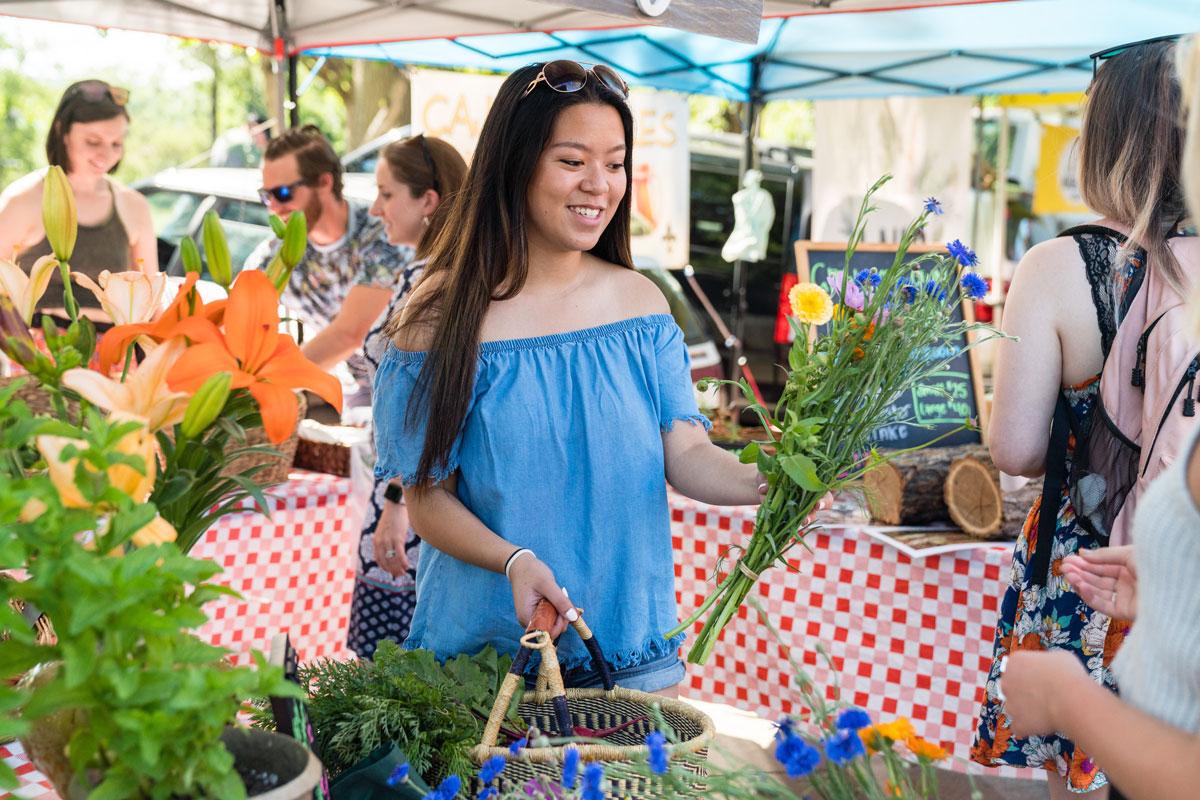 A woman holds a basket of produce and picks up a bunch of flowers at a stand within the farmers market