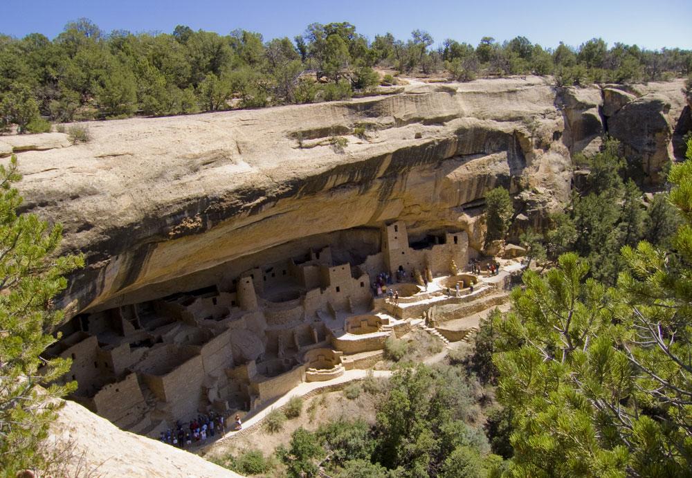 Brick towers and kivas sit on a ledge beneath a cliff wall that's topped by green trees