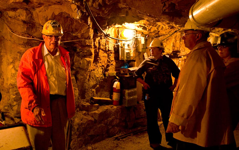 A tour guide in a red jacket and yellow hard hat leads a group of people through a dimly lit tunnel