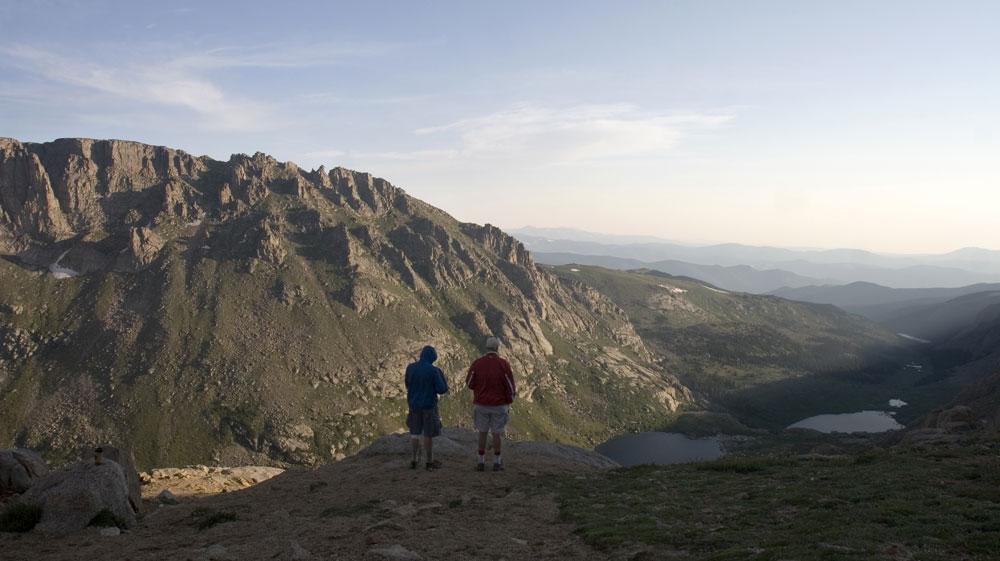 Two people atop Mount Blue Sky gaze onto the light-blue horizon, surrounded by the tops of shorter mountains