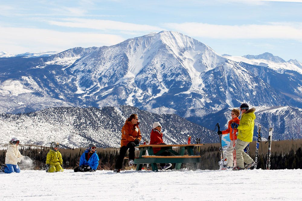 At a picnic table on a snowy mesa, a group of kids and adults gather for lunch. One giant, snow-covered peak rises behind them