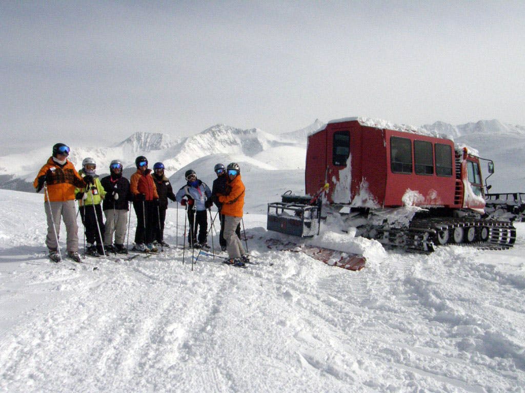 A red snowcat lets its passengers out at the top of a backcountry ski slope. They all pose for the camera before heading down the mountain.