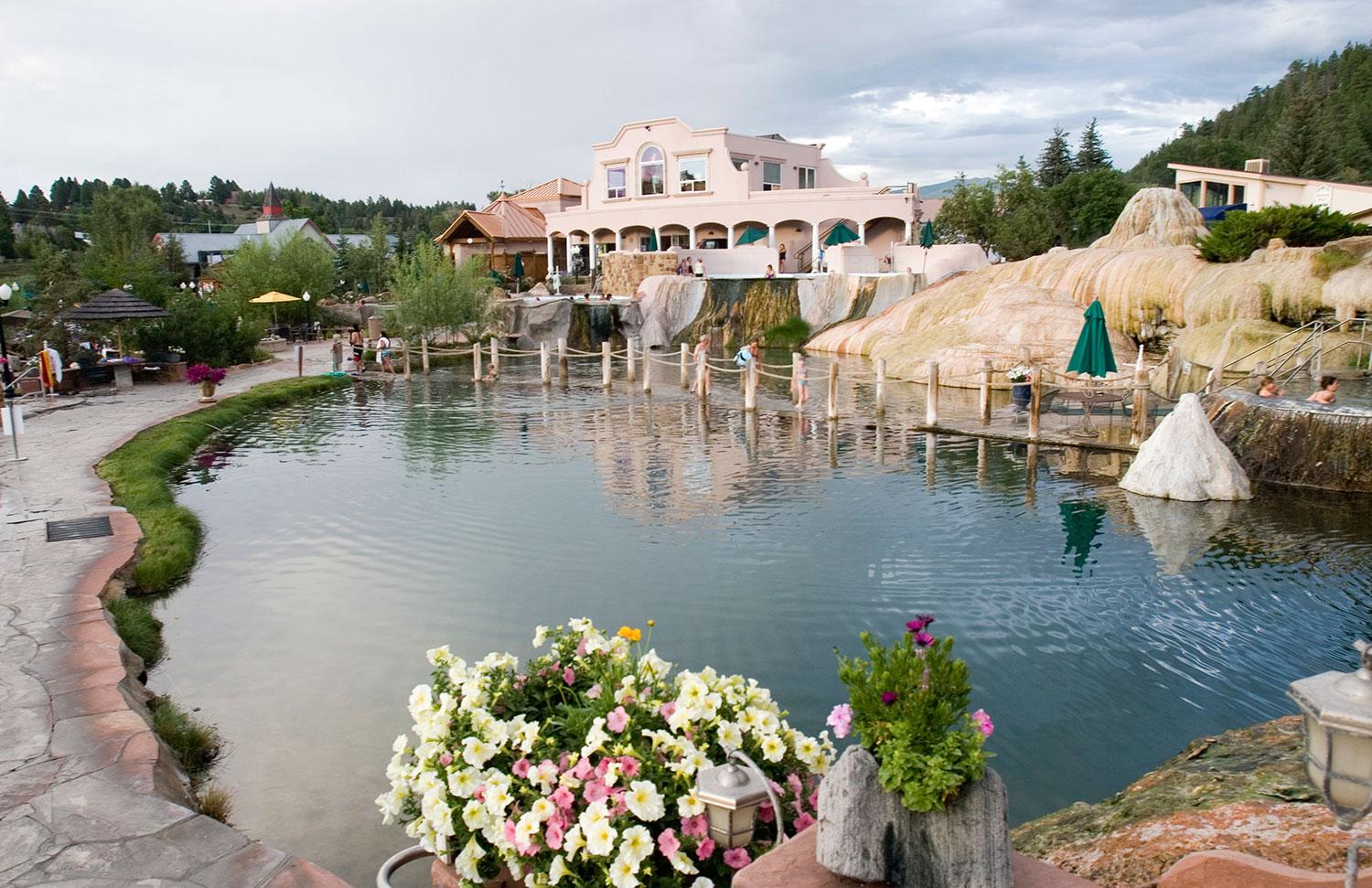 Flowers grow near the edge of The Springs Resort and Spa hot springs. The pool is rippling slightly with a two story building in the distance beneath a grey sky.