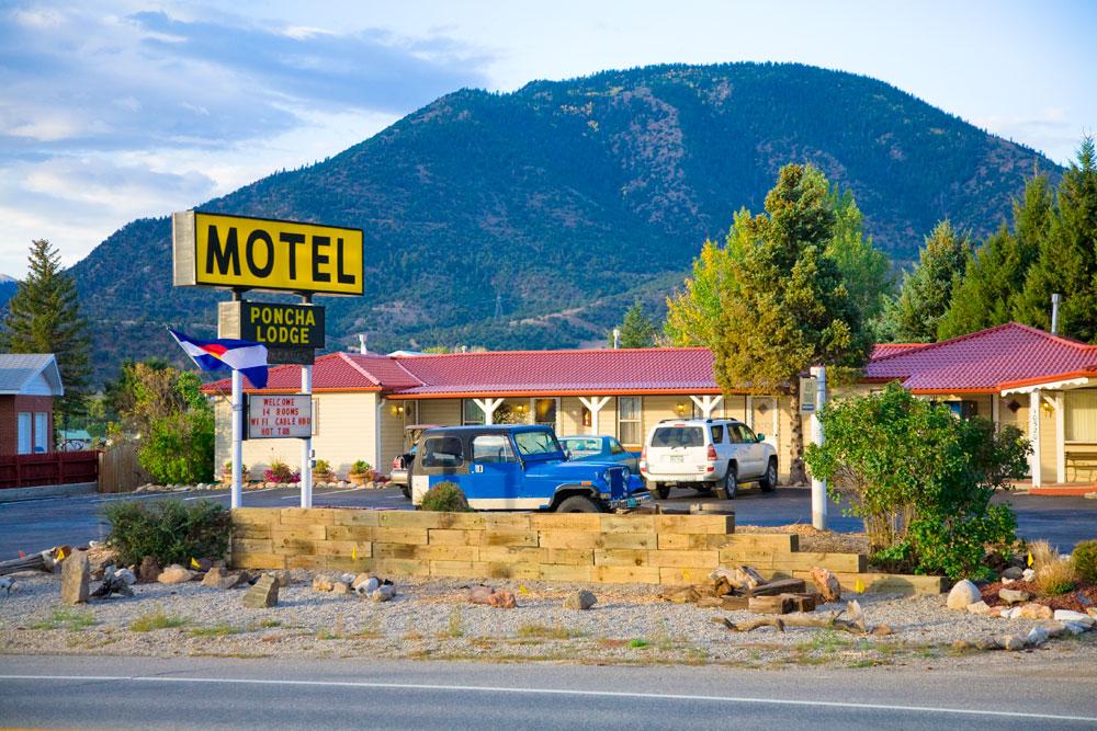 The "Motel Poncha Lodge" sits under a tree-covered mountain. The motel has a red roof with yellow walls and multiple cars in the parking lot.