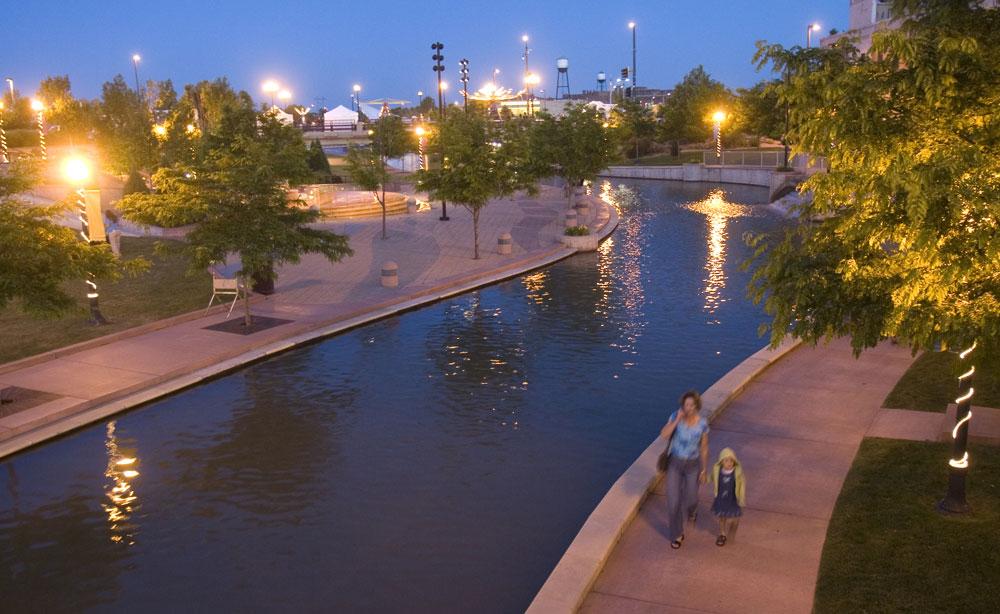 A brown river winds between a red-brick riverwalk lined with trees and streetlights