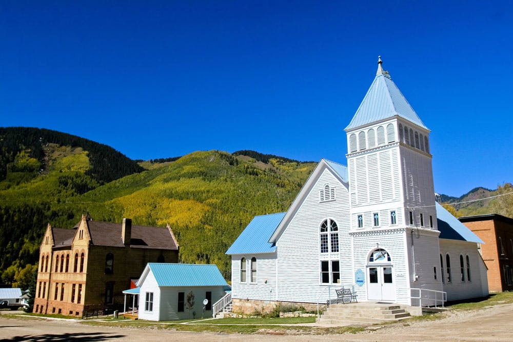 A historic blue-roofed chapel sits next to other historical buildings beneath a deep-blue sky with evergreen-covered mountains.