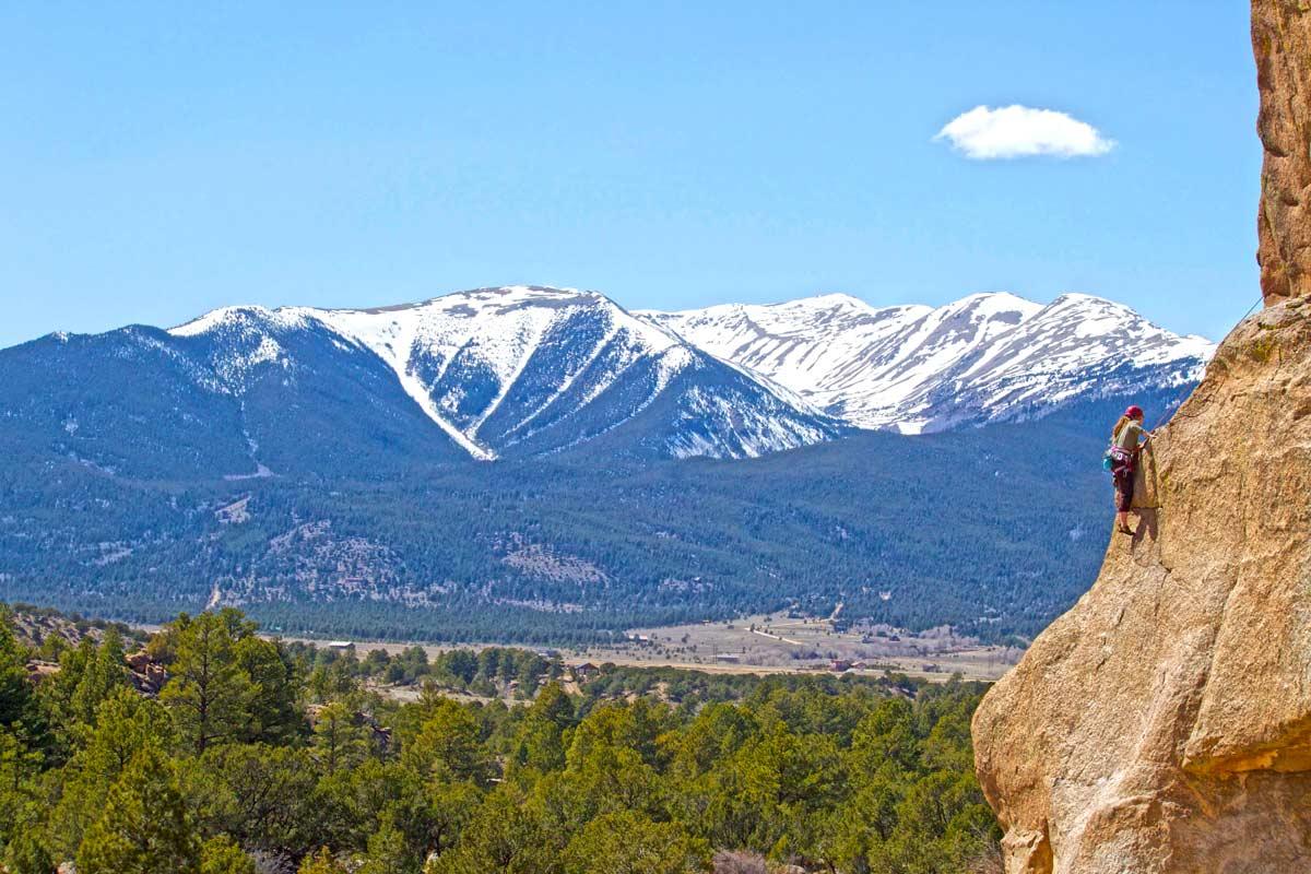 A woman rock climbs on the far right of the image with snow-covered mountains in the background. There's a blue sky with one whispy cloud and there are evergreen trees. 