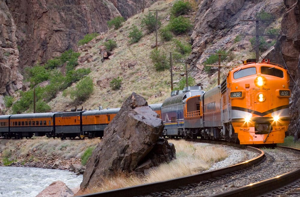 A pumpkin-orange train engine chugs along the curve of a track near Cañon City, Colorado. It pulls several passenger cars behind it.