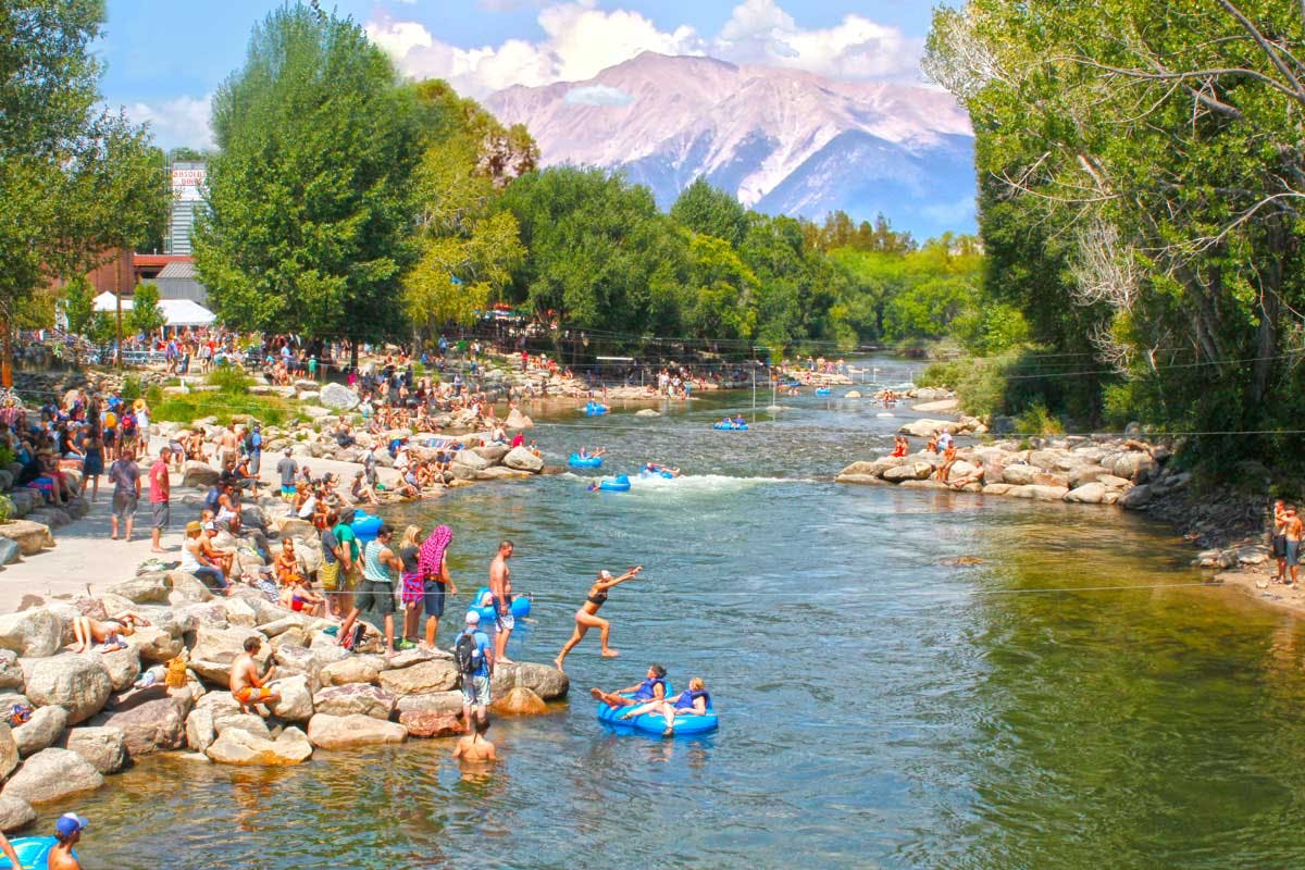 People wait to jump into the river on a blue-sky summer's day in Salida. There's a mountain in the distance and green trees thrive on both riverbanks.