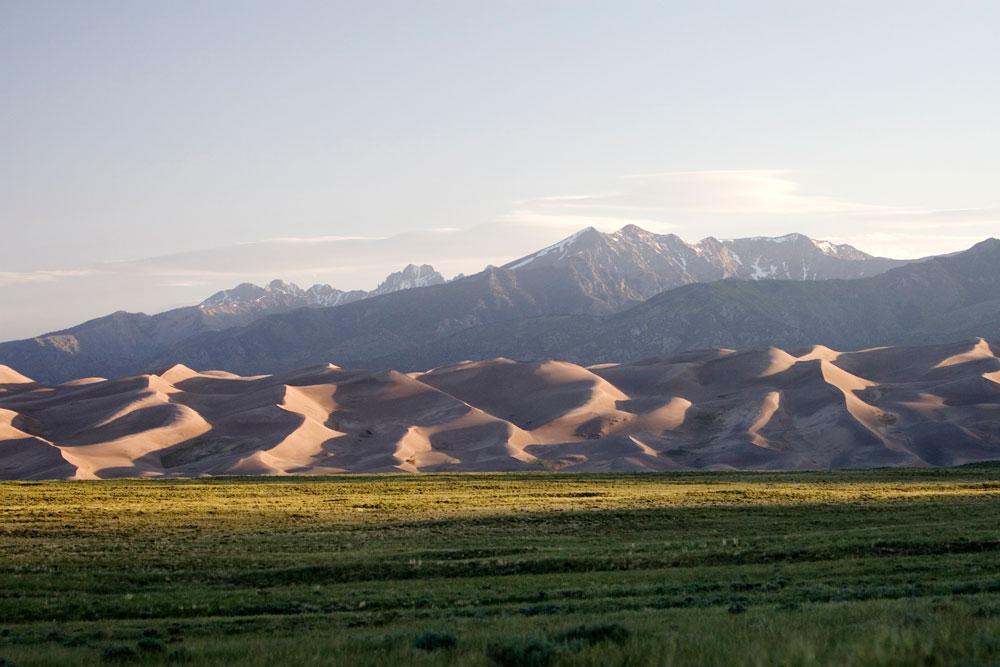 A field of low grass lays before the wavy sand dunes and tower, snow-capped peaks of Great Sand Dunes National Park and Preserve near Alamosa.