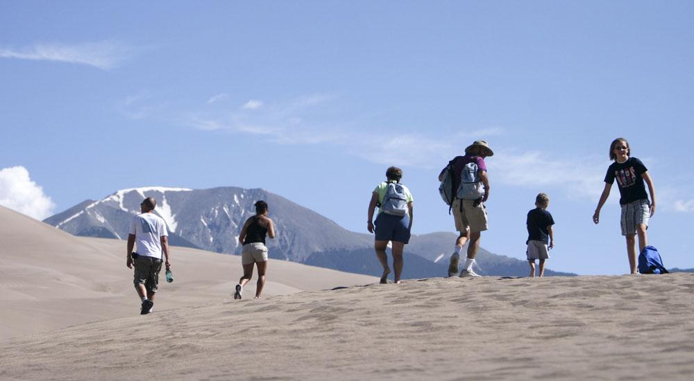 Six people walk through the dune field wearing hats under the bright sun; the wind blows as they make their way