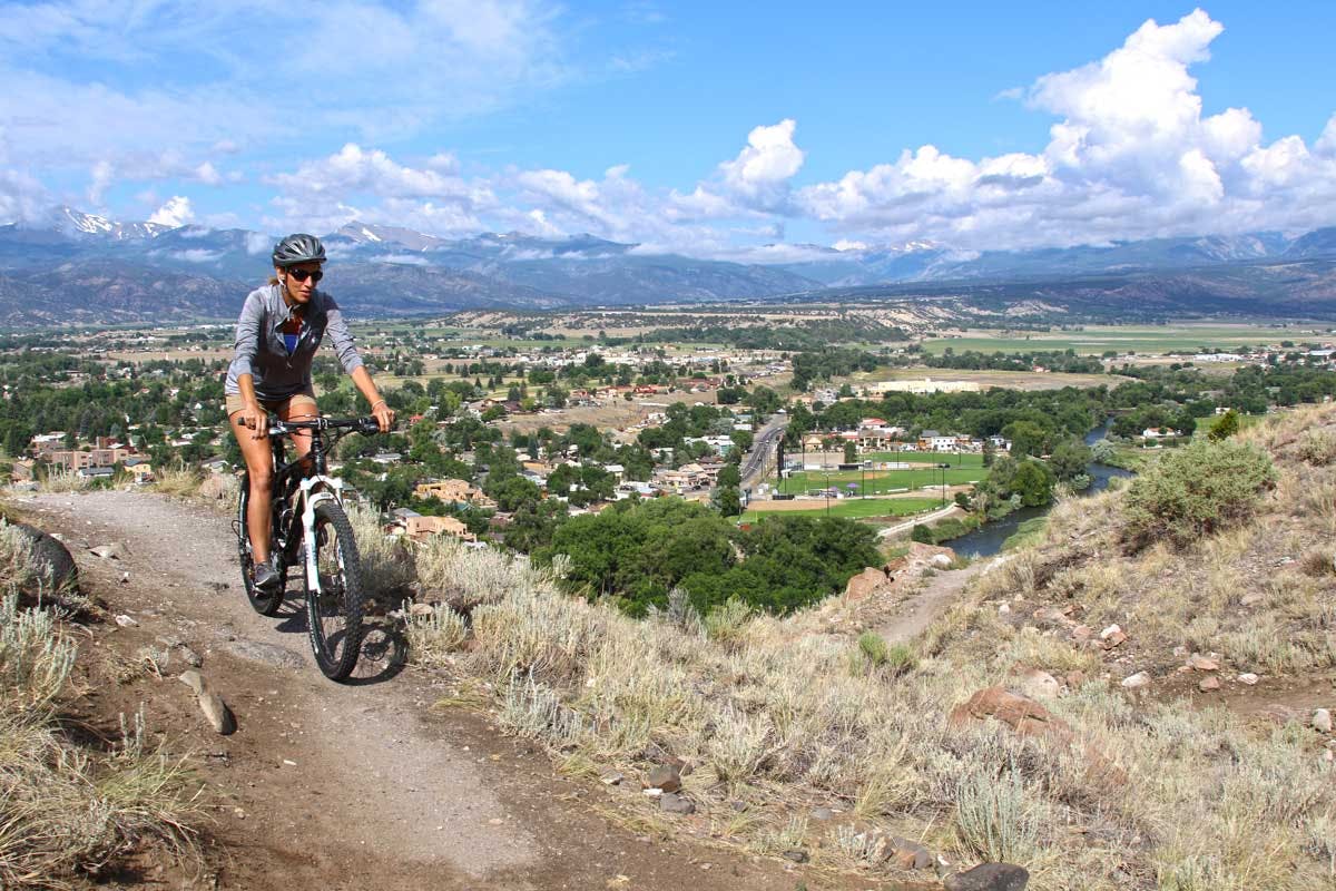 A person mountain bikes on a thin path along a ridge in the summer. The town of Salida sits below with green grasses and trees below a blue sky.