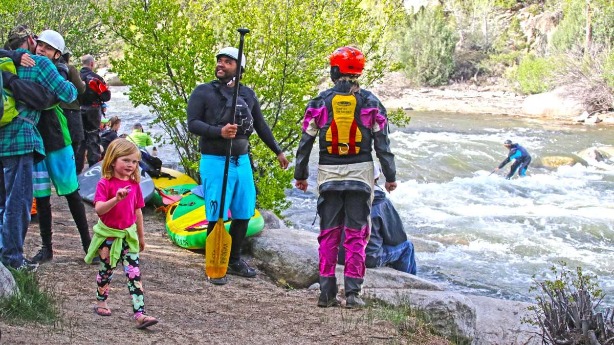 People and one child stand on the riverbank watching a person whitewater kayak at Buena Vista Whitewater Park in the summer.