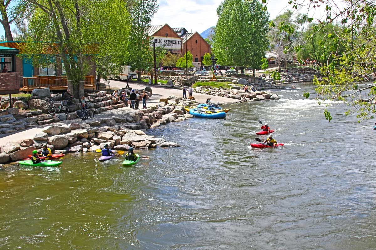 Six kayakers sit in the Arkansas River in downtown Salida. There are trees with green leaves and the bank of the river has boulders.